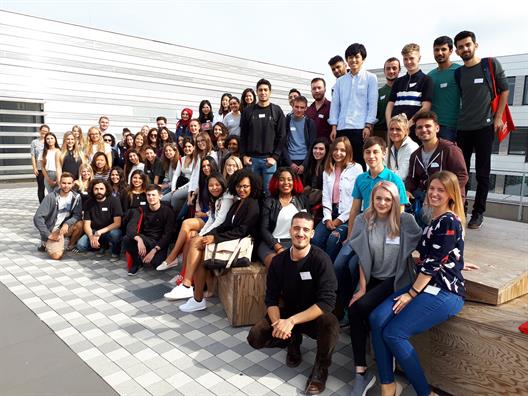 a big Group of Young students is smiling into the camera, behind them is a Grey Building and blue sky.
